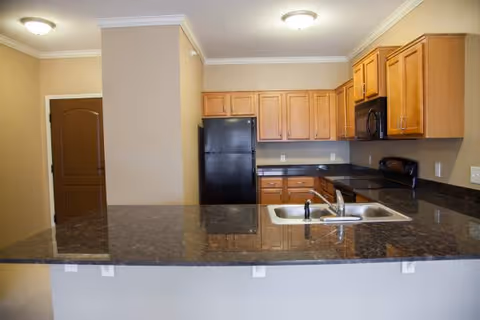 Interior view of a kitchen in a senior living apartment featuring wooden cabinets, a black refrigerator, a black microwave, a black stove, and a double sink set in a dark granite countertop with beige walls and ceiling lights.