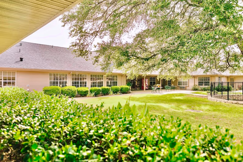 A bright outdoor view of a single-story building with large windows, surrounded by green bushes and a well-maintained lawn. There is a large tree providing shade over a patio area with tables and chairs, and a vending machine is visible near the building entrance.