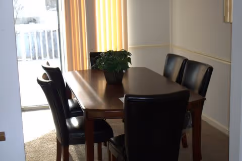 A dining area with a rectangular wooden table surrounded by six black leather chairs. A green potted plant is placed in the center of the table. Vertical blinds cover a window or sliding door in the background, allowing natural light to enter the room.