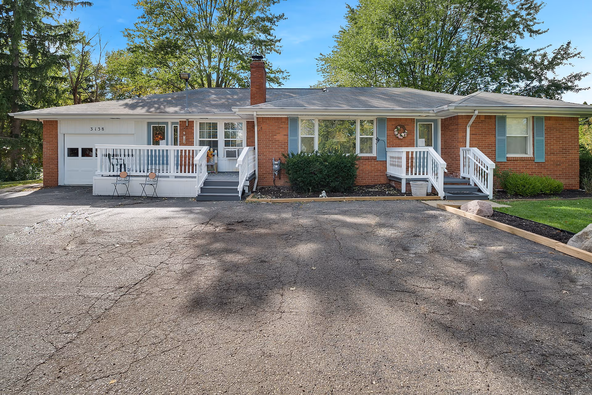 Front exterior view of a single-story brick building with a gray roof, white railings, and blue shutters. There are two small porches with steps leading up to the entrances, a garage door on the left side, and some outdoor chairs on the porch. Trees and greenery surround the building under a clear blue sky.