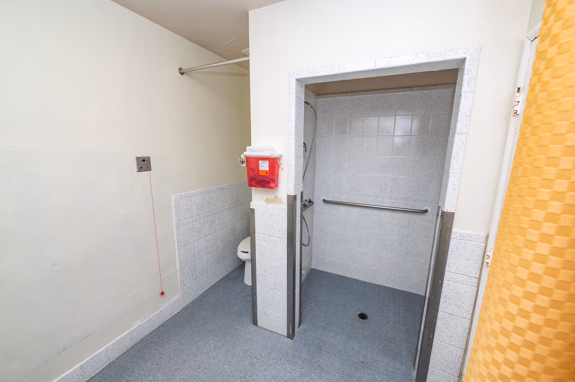 A bathroom with an accessible shower area featuring a grab bar and handheld showerhead. There is a toilet partially visible behind a half wall, a red emergency pull cord on the wall, and a red sharps disposal container mounted on the wall.