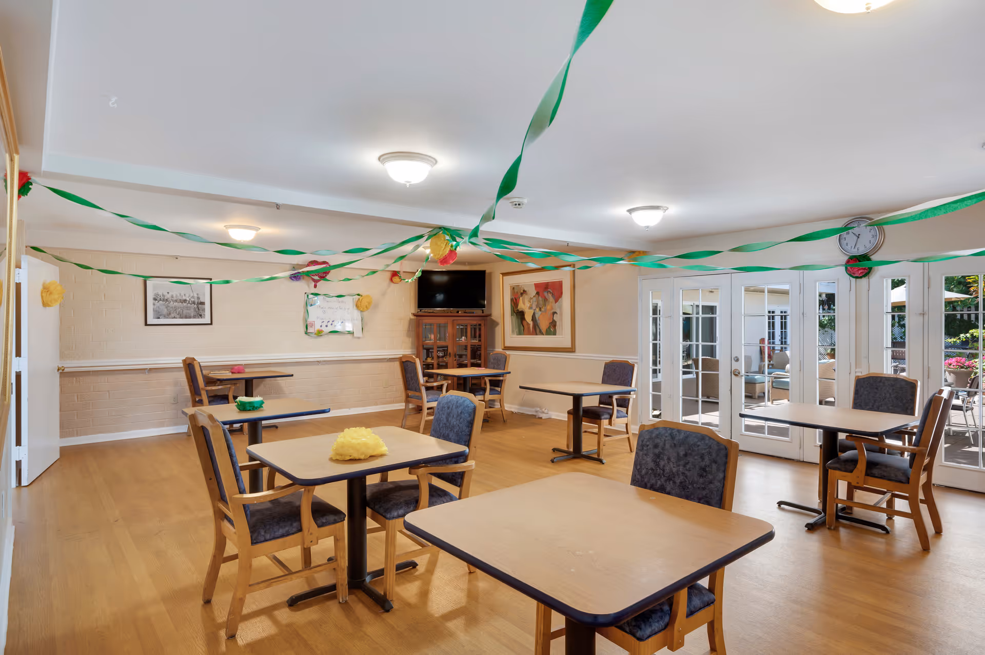 Bright communal dining/activity room with several tables and chairs, green streamers hanging from the ceiling, and French doors leading outside.