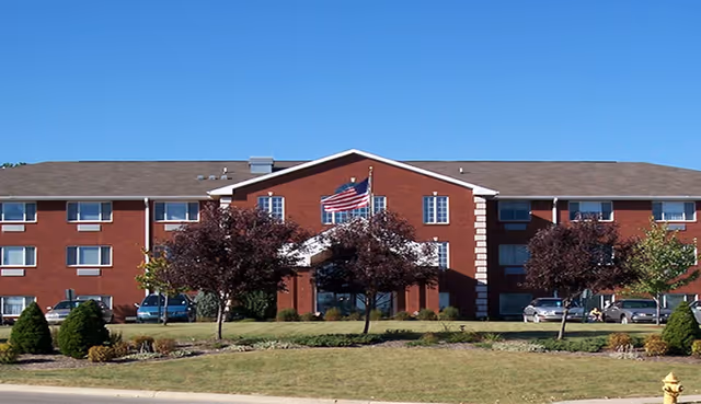 Red-brick, three-story senior living building with an American flag and landscaped front lawn under a clear blue sky.