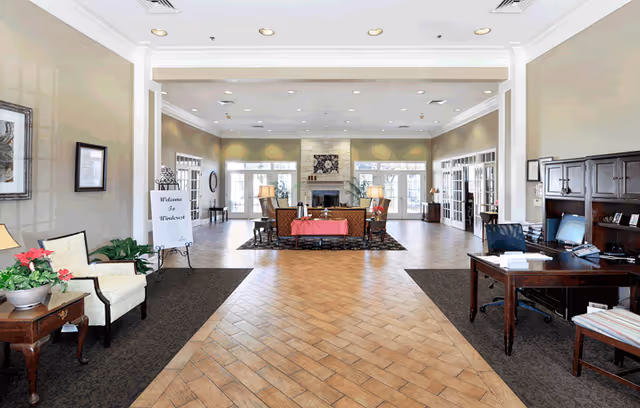 A spacious and well-lit common area with a tiled floor and high ceiling. The room features a seating area with sofas and a coffee table centered on a rug in front of a stone fireplace. There are multiple windows and glass doors letting in natural light. On the left side, there is a small table with a potted plant and a chair, and a sign on an easel that reads 'Welcome to Windsor'. On the right side, there is a wooden desk with a computer and office chair. The walls are painted beige and decorated with framed artwork.