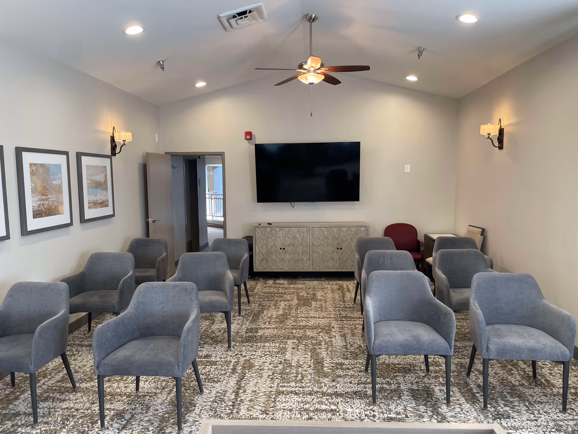 A small meeting or presentation room with multiple gray upholstered chairs arranged in rows facing a wall-mounted flat screen TV. The room has a patterned carpet, light gray walls with three framed artworks on the left wall, two wall sconces with lampshades, a ceiling fan with a light, and a cabinet beneath the TV. There is an open door on the left side leading to another area.