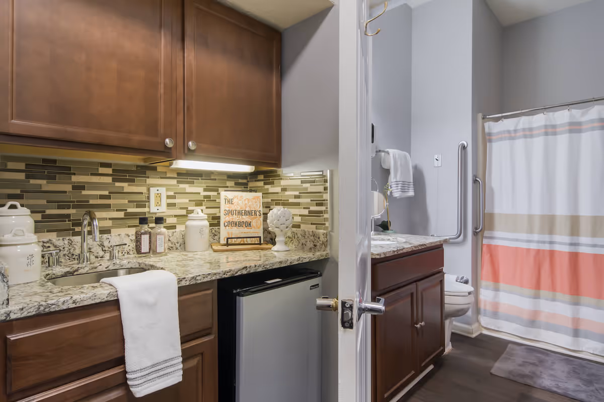 A kitchenette area with a granite countertop, brown cabinets, a small sink, and a mini refrigerator. To the right, there is a bathroom with a shower curtain featuring horizontal stripes in white, beige, and coral colors, a toilet, and a vanity with a granite countertop and brown cabinets. The bathroom has grab bars installed near the toilet and shower.
