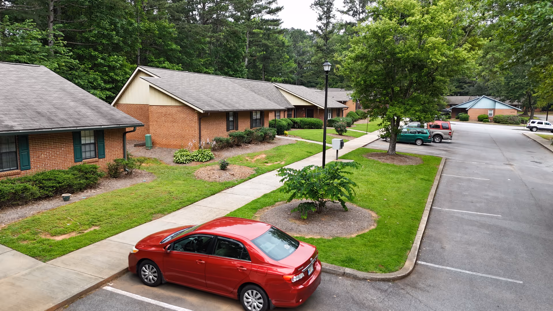 View of a senior living facility parking area with several single-story brick buildings surrounded by green lawns, trees, and shrubs. A red car is parked in the foreground along a sidewalk that runs between the buildings and the parking lot.