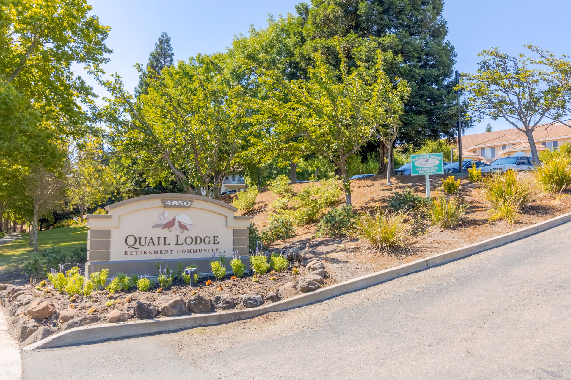 Entrance sign reading 'Quail Lodge Retirement Community' surrounded by landscaped plants and trees with buildings visible behind.