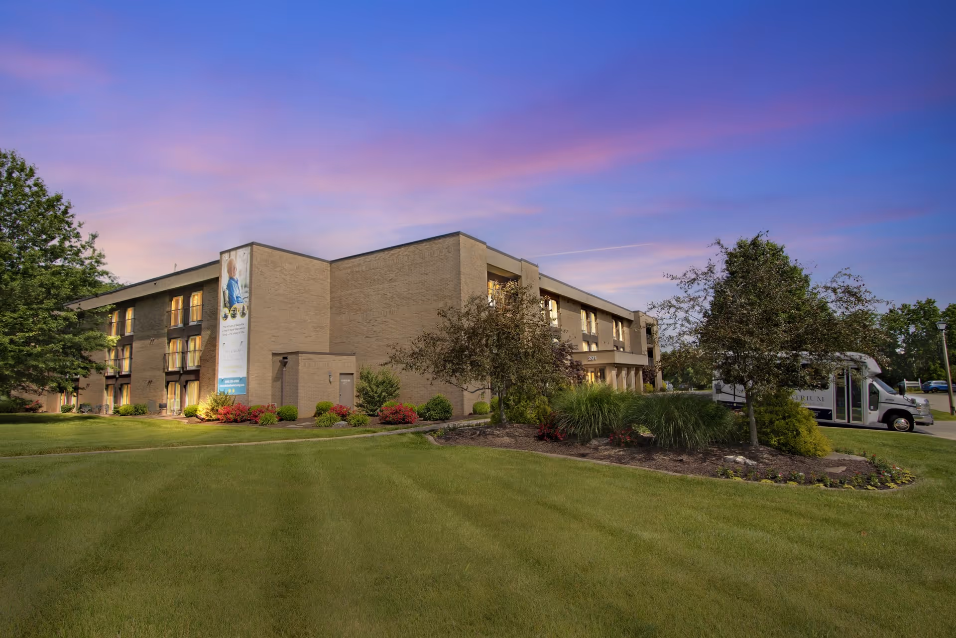 Three-story brick senior living building with landscaped lawn and a shuttle van under a colorful evening sky.