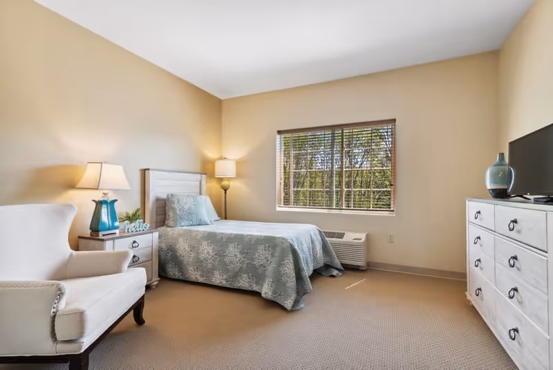 A cozy bedroom with a single bed covered in a blue patterned bedspread, a white armchair, a wooden nightstand with a lamp and decorative items, a window with blinds showing trees outside, a floor lamp in the corner, and a white dresser with a TV and a vase on top.