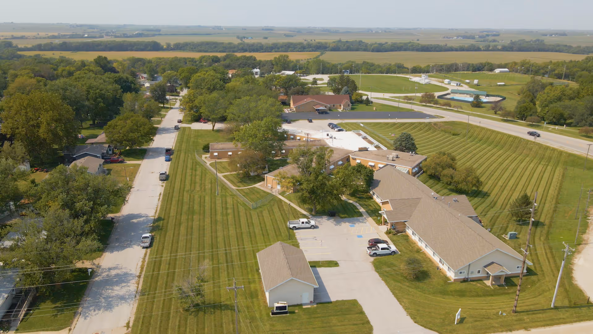 Aerial view of Oakland Manor senior living facility showing multiple single-story buildings with beige roofs surrounded by well-maintained lawns and trees. There is a parking lot with several cars, a road running alongside the property, and open fields in the background under a clear sky.