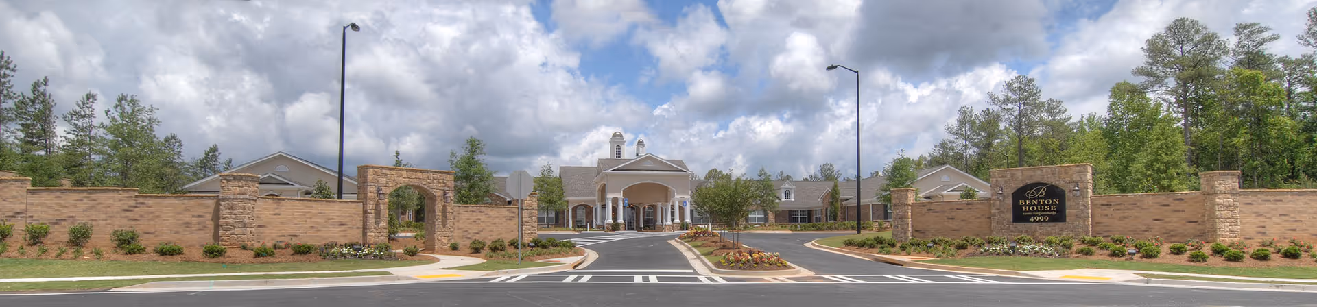 Wide panoramic view of the entrance to Benton House of Douglasville, showing a large brick wall with an arched gateway and a sign with the facility's name. The driveway leads to a covered entrance of a single-story building surrounded by trees and landscaping under a partly cloudy sky.