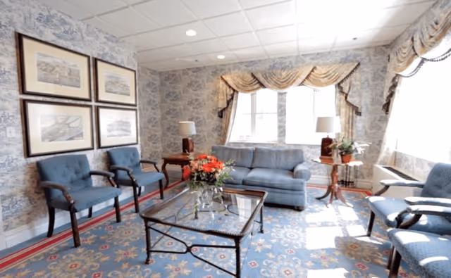 A bright living room with blue upholstered chairs and a matching sofa arranged around a glass coffee table with a flower arrangement. The room features patterned wallpaper, framed artwork on the wall, two table lamps on side tables, and large windows with decorative curtains allowing natural light to fill the space.