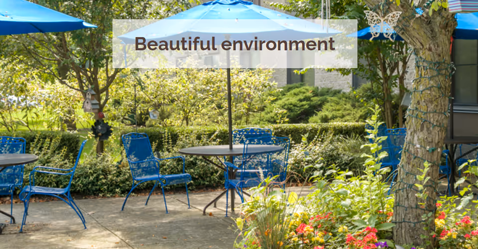 Outdoor patio area with blue metal chairs and tables under blue umbrellas surrounded by greenery, trees, and colorful flowers. A translucent banner with the text 'Beautiful environment' is displayed in the center.