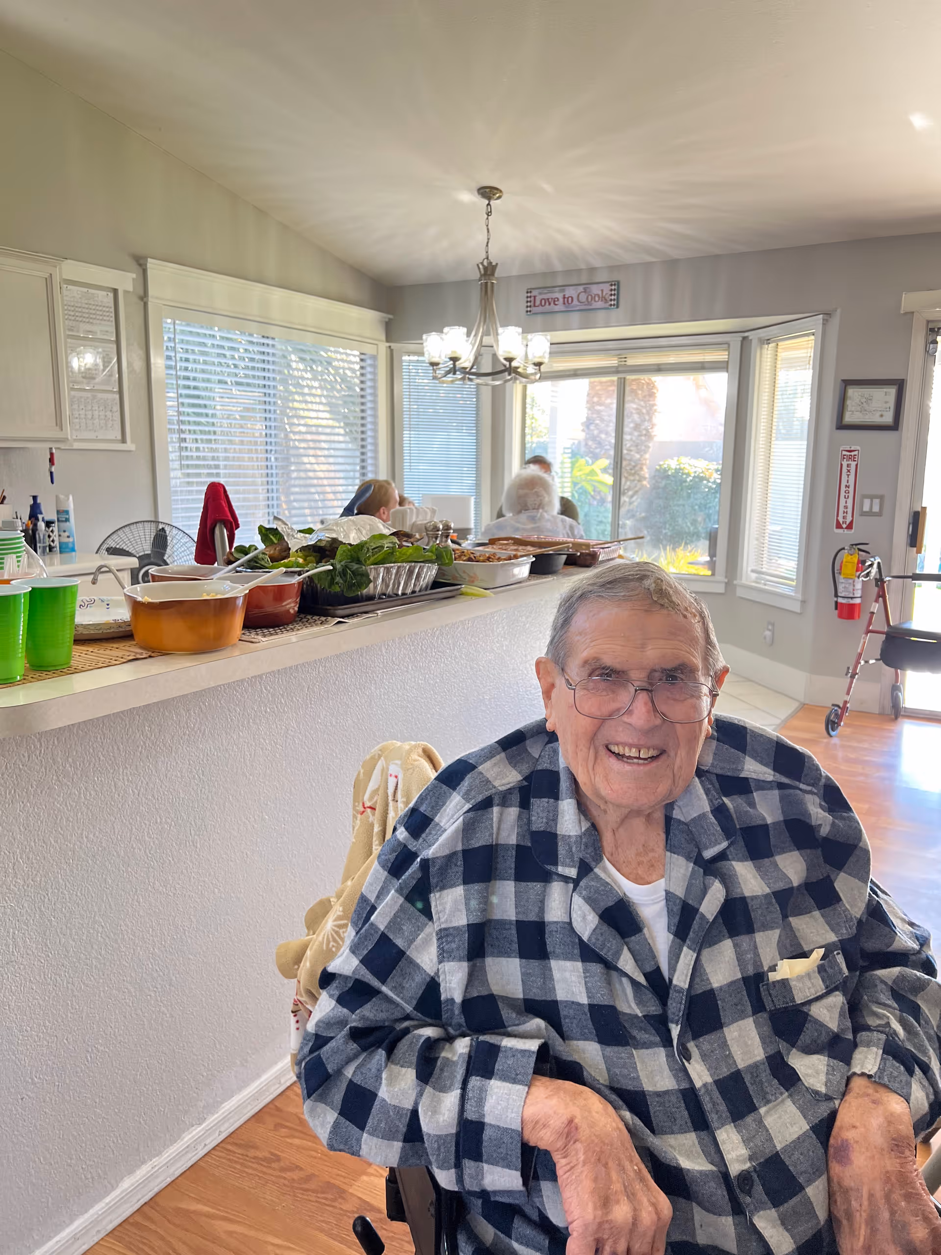 An elderly man wearing glasses and a checkered shirt is sitting in a wheelchair and smiling in a bright room with large windows. Behind him, there is a counter with various food dishes and green cups. In the background, two other elderly individuals are seated near the windows. The room has light-colored walls, wooden flooring, and a chandelier hanging from the ceiling. A sign above the window reads 'Love to Cook'.
