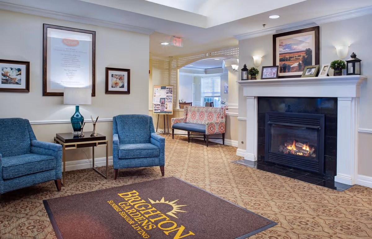 A cozy senior living facility common area with two blue armchairs and a side table with a lamp. There is a fireplace with a lit fire on the right side, decorated with framed pictures, plants, and lanterns on the mantel. A patterned bench is visible in the background near a bulletin board. The floor has a patterned carpet with a large mat that reads 'Brighton Gardens Sunrise Senior Living'.