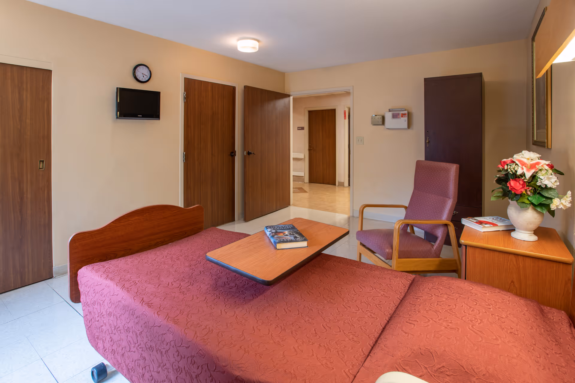 A senior living facility bedroom with a bed covered in a red patterned bedspread, a wooden side table with a vase of flowers and books, a purple armchair, a wall-mounted TV, and several wooden doors leading to other rooms or hallways.