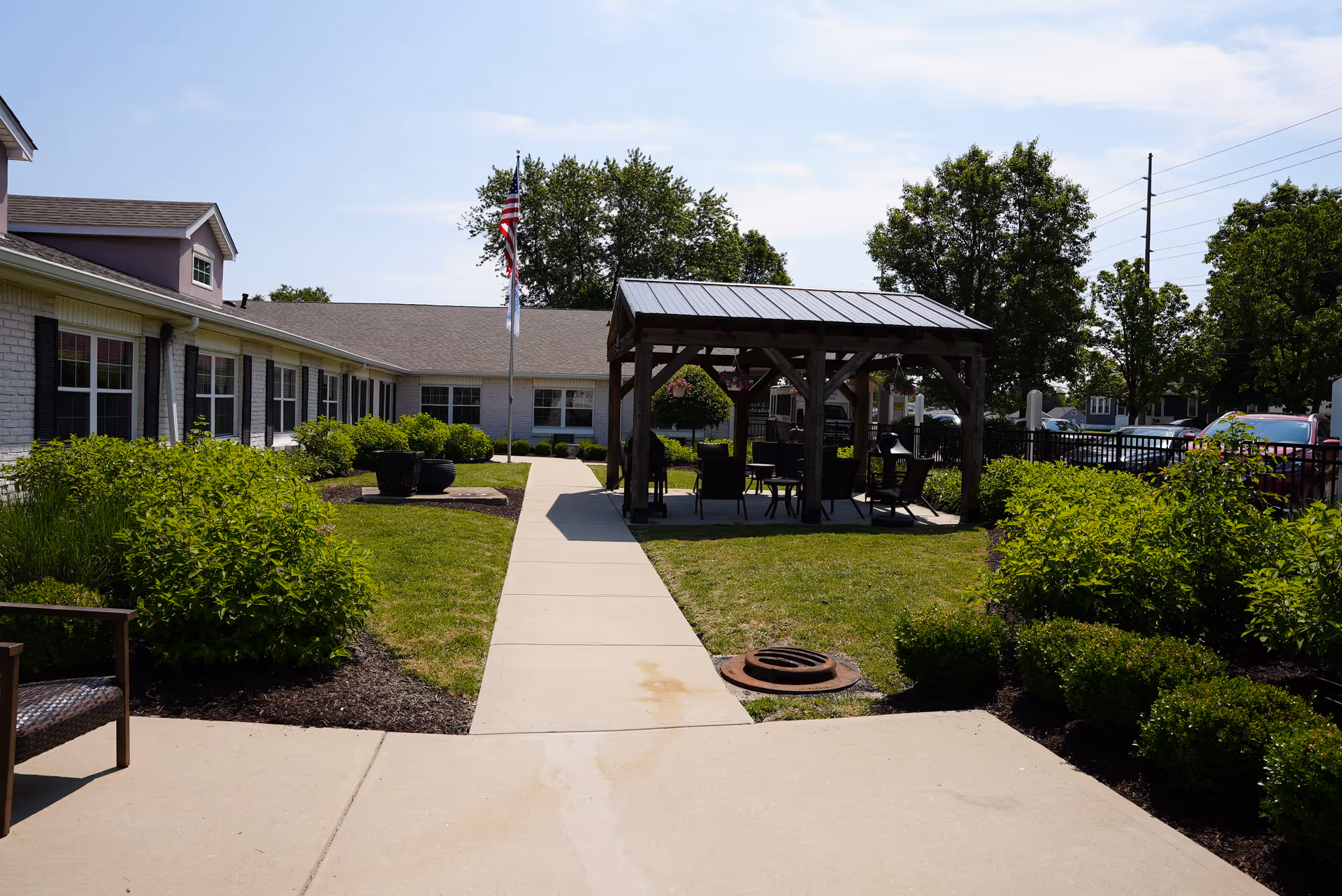 Courtyard with a paved walkway leading to a wooden gazebo surrounded by lawn, shrubs, a flagpole, and a low building under a clear sky.