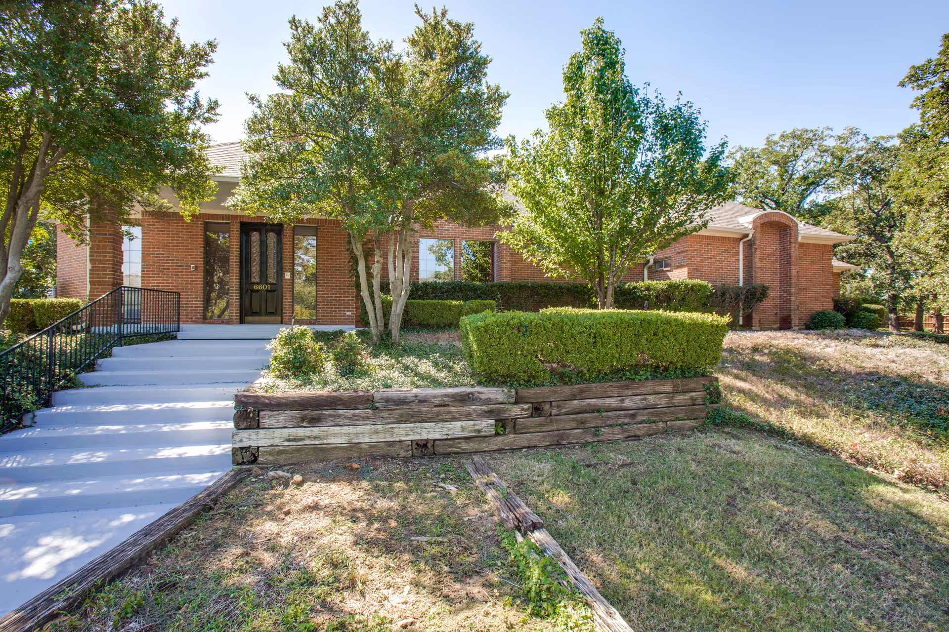 Front exterior of a brick memory care building with steps and a walkway leading to a dark entry door, surrounded by trees, hedges, and a wooden retaining wall.