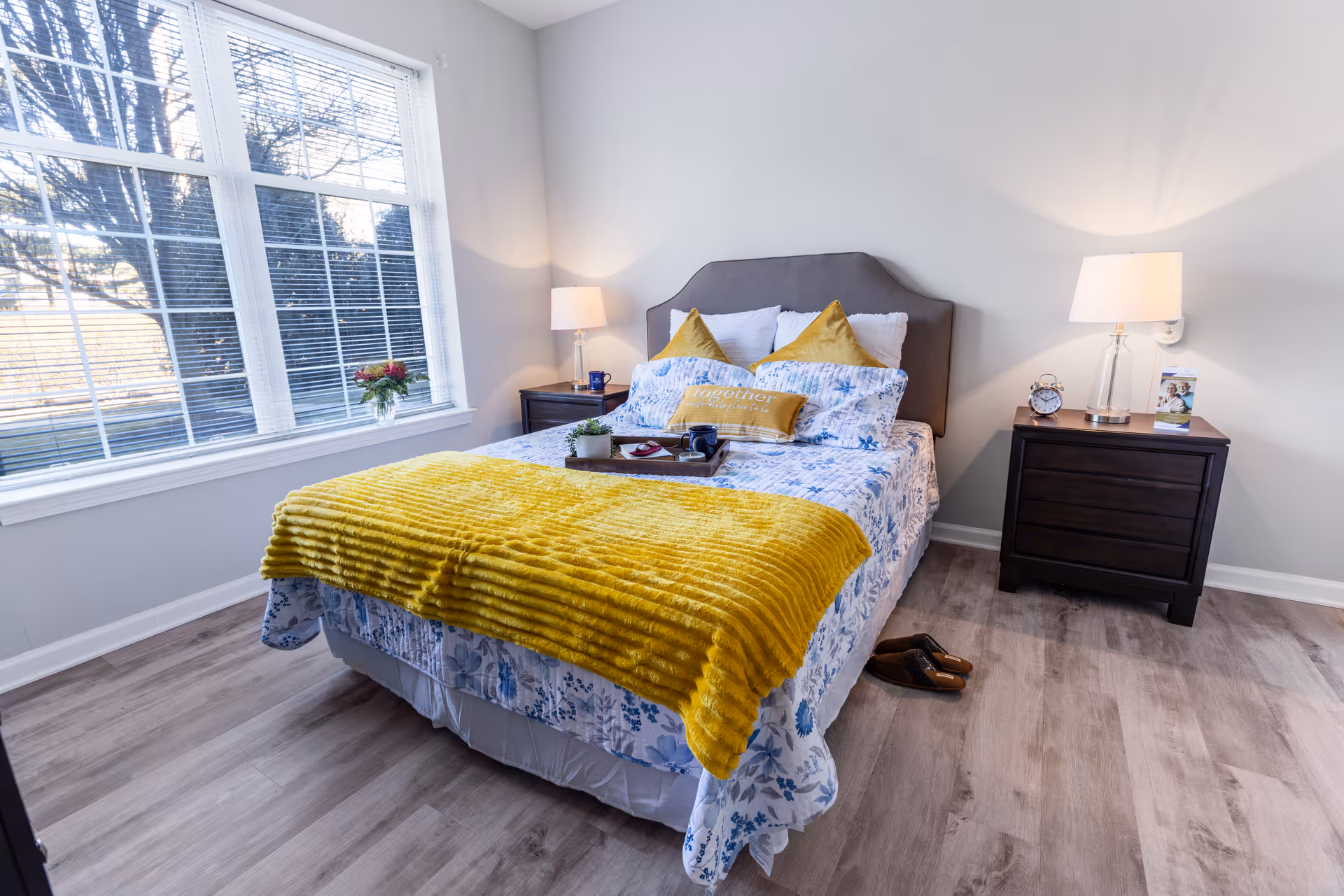 A bright bedroom with a large window on the left letting in natural light. The bed has a gray upholstered headboard, white and yellow pillows, a floral bedspread, and a yellow throw blanket. There are two dark wooden nightstands on either side of the bed, each with a lamp. One nightstand also has an alarm clock and a brochure. A pair of brown slippers is on the floor next to the bed.
