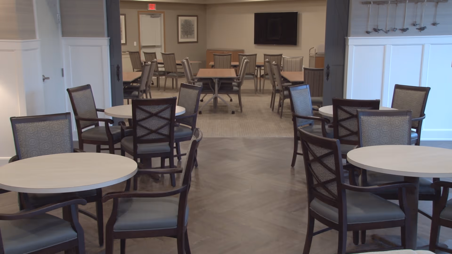 A spacious dining area in a senior living facility with multiple round and rectangular tables surrounded by cushioned chairs. The room has neutral-colored walls, a wooden floor, and a large flat-screen TV mounted on the far wall. There is a door and framed artwork visible in the background.