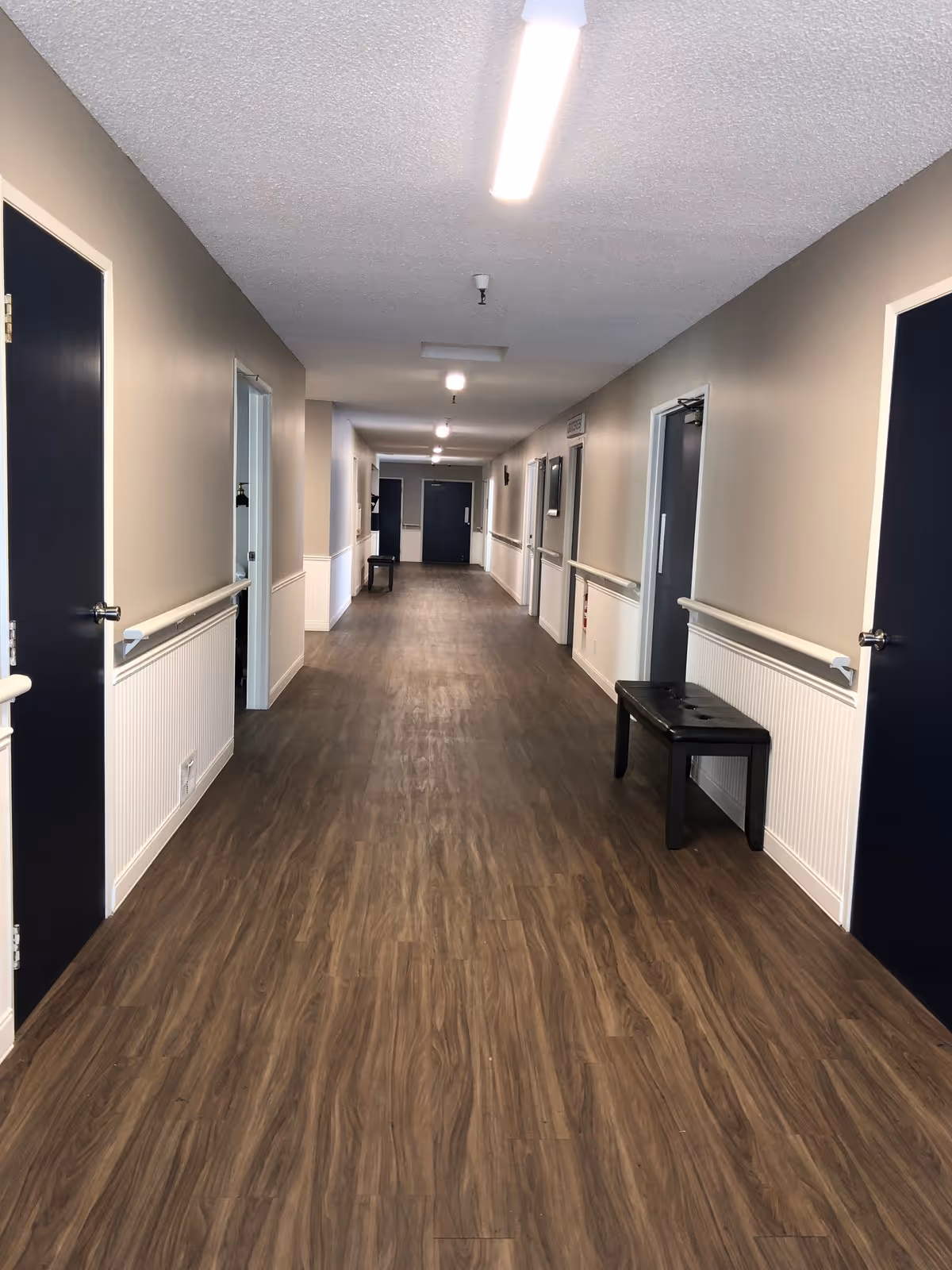 A long indoor hallway with wood-patterned flooring, beige walls with white wainscoting, and dark blue doors on both sides. There are handrails along the walls and a few benches placed against the walls. The ceiling has several light fixtures providing illumination.