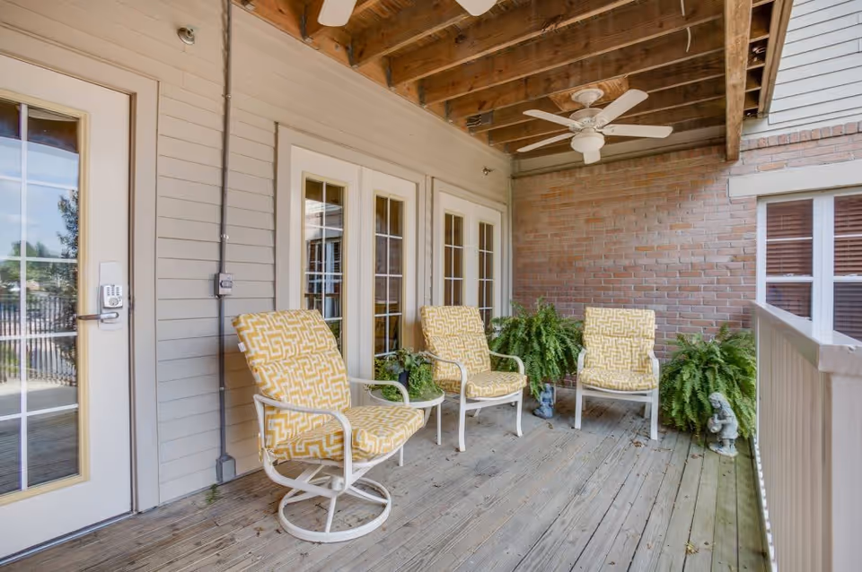 Covered outdoor patio area with three cushioned chairs featuring yellow patterned upholstery, two potted ferns, small decorative statues, wooden deck flooring, and a ceiling fan under exposed wooden beams. French doors and a door with a keypad lock lead inside the building.