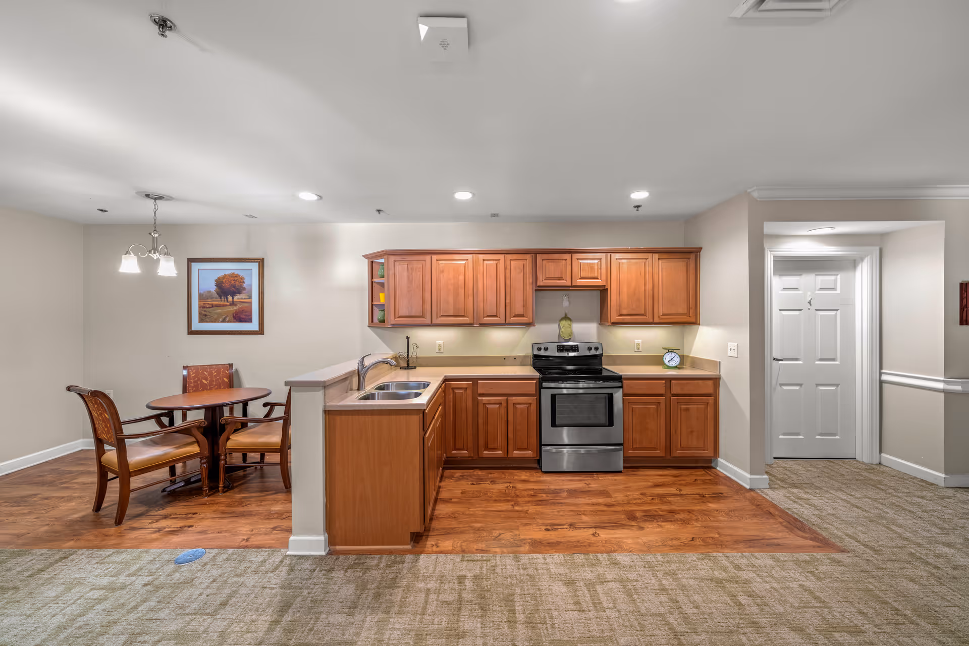 Interior view of a senior living facility kitchen and dining area. The kitchen features wooden cabinets, a stainless steel stove, and a double sink. Adjacent to the kitchen is a small dining area with a round wooden table and four chairs. The walls are light-colored, and there is a framed picture hanging above the dining table. The floor transitions from carpet in the foreground to wood flooring in the kitchen and dining area.