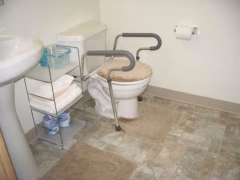 A bathroom with a toilet featuring grab bars, sink, and shelf unit.
