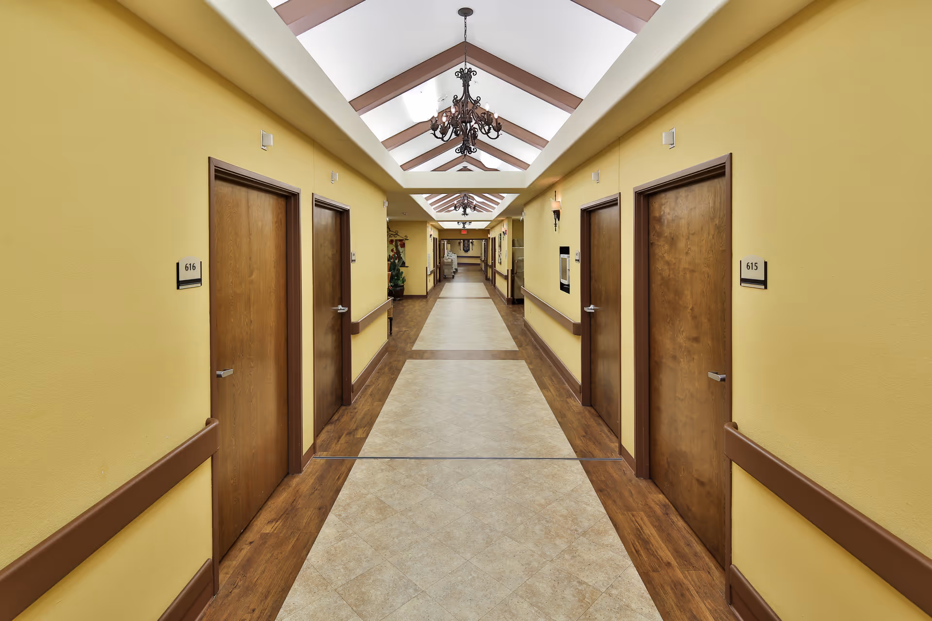 A long, well-lit hallway in a senior living facility with yellow walls, wooden doors on both sides labeled with room numbers 615 and 616, and a tiled floor with wooden borders. The ceiling features skylights and decorative chandeliers.