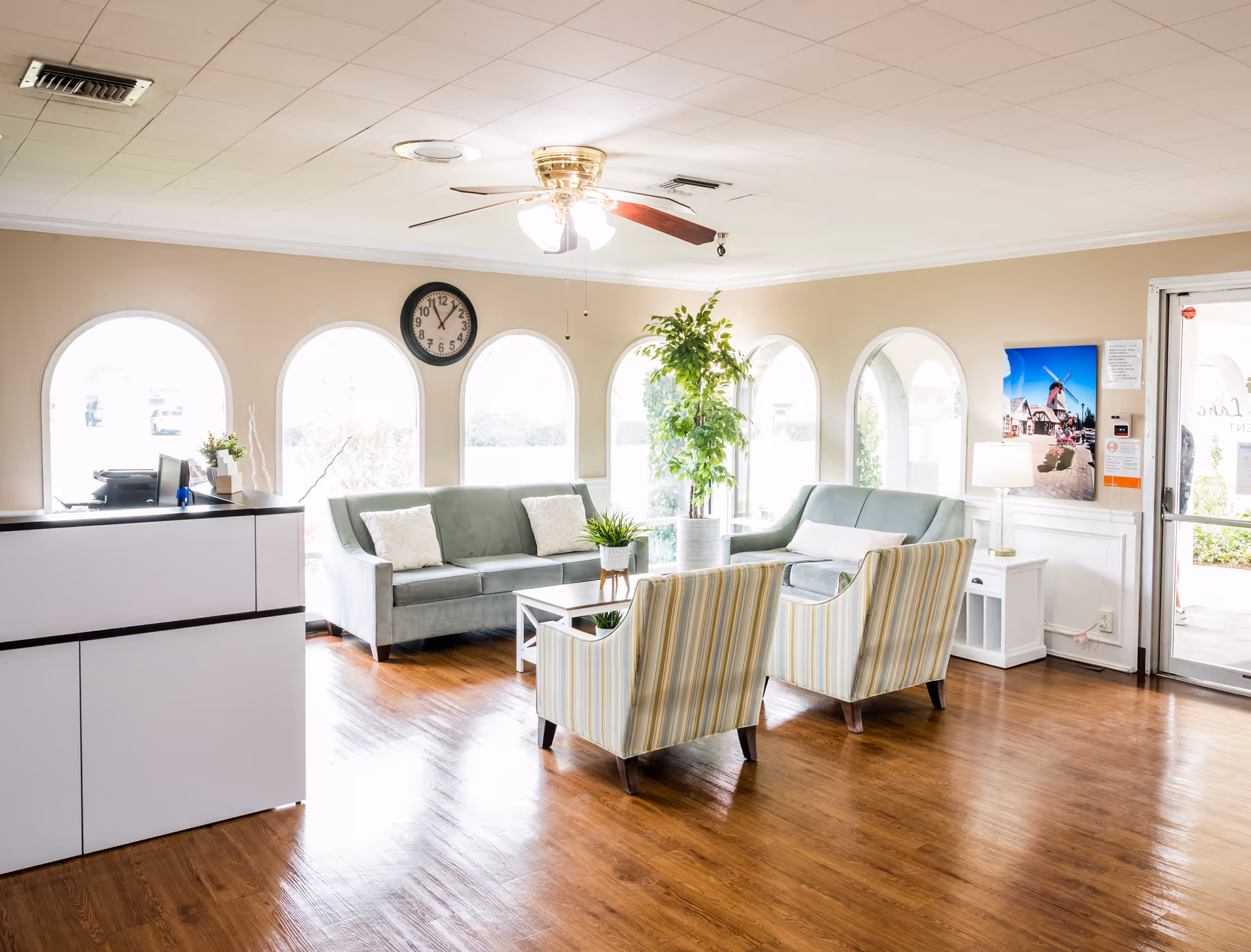 A bright and welcoming reception area with a white reception desk on the left, two green sofas, two striped armchairs, a coffee table with a plant, a tall potted plant, a ceiling fan with lights, and arched windows letting in natural light. A clock is mounted on the wall above the sofas, and a door with glass panels is visible on the right side.
