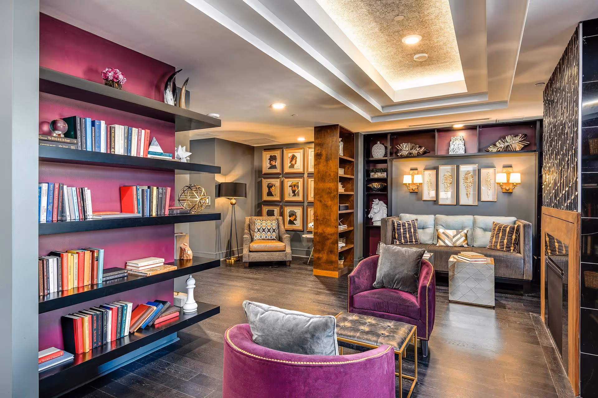 A cozy and stylish living room area with dark wood flooring, a purple accent wall with built-in black shelves filled with books and decorative items, two purple armchairs with gray cushions, a tufted bench, a gray sofa with patterned pillows, framed artwork on the walls, and warm lighting from wall sconces and ceiling lights.