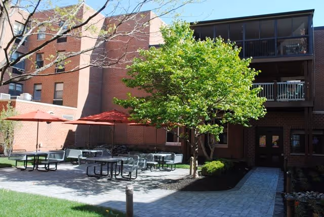 Outdoor patio area at Philadelphia Protestant Home with several picnic tables and red umbrellas, surrounded by a brick building and green trees under a clear sky.