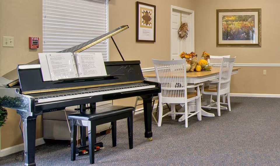 A common room featuring a black grand piano beside a dining table with white chairs and autumn centerpiece decorations.