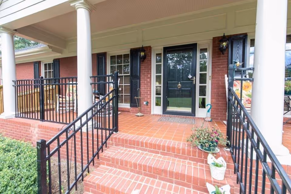 Front porch of a brick building with white columns, black railings, and a black front door with glass panels on either side. There are potted plants on the steps and a rocking chair on the porch.