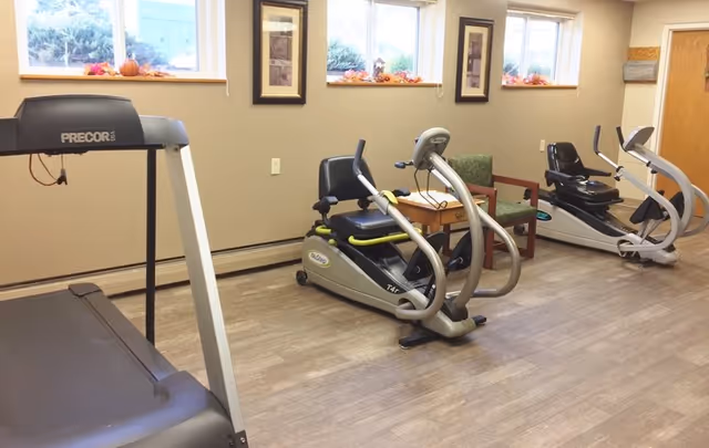 A small exercise room with a treadmill, two recumbent exercise bikes, a wooden side table, and a green upholstered chair. The room has light-colored walls, three windows with fall-themed decorations on the sills, and framed artwork on the walls.