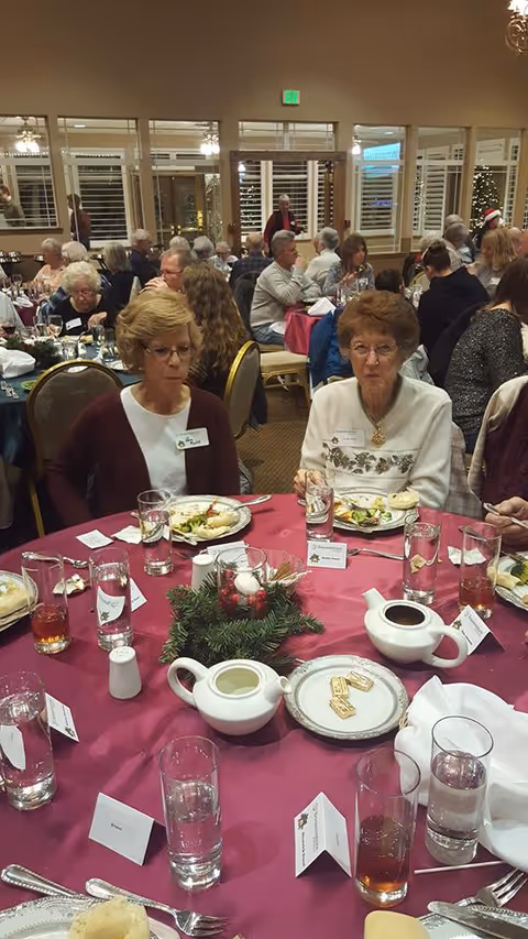 A dining room with seniors seated at round banquet tables, two elderly women in the foreground eating amid place settings and a holiday centerpiece.