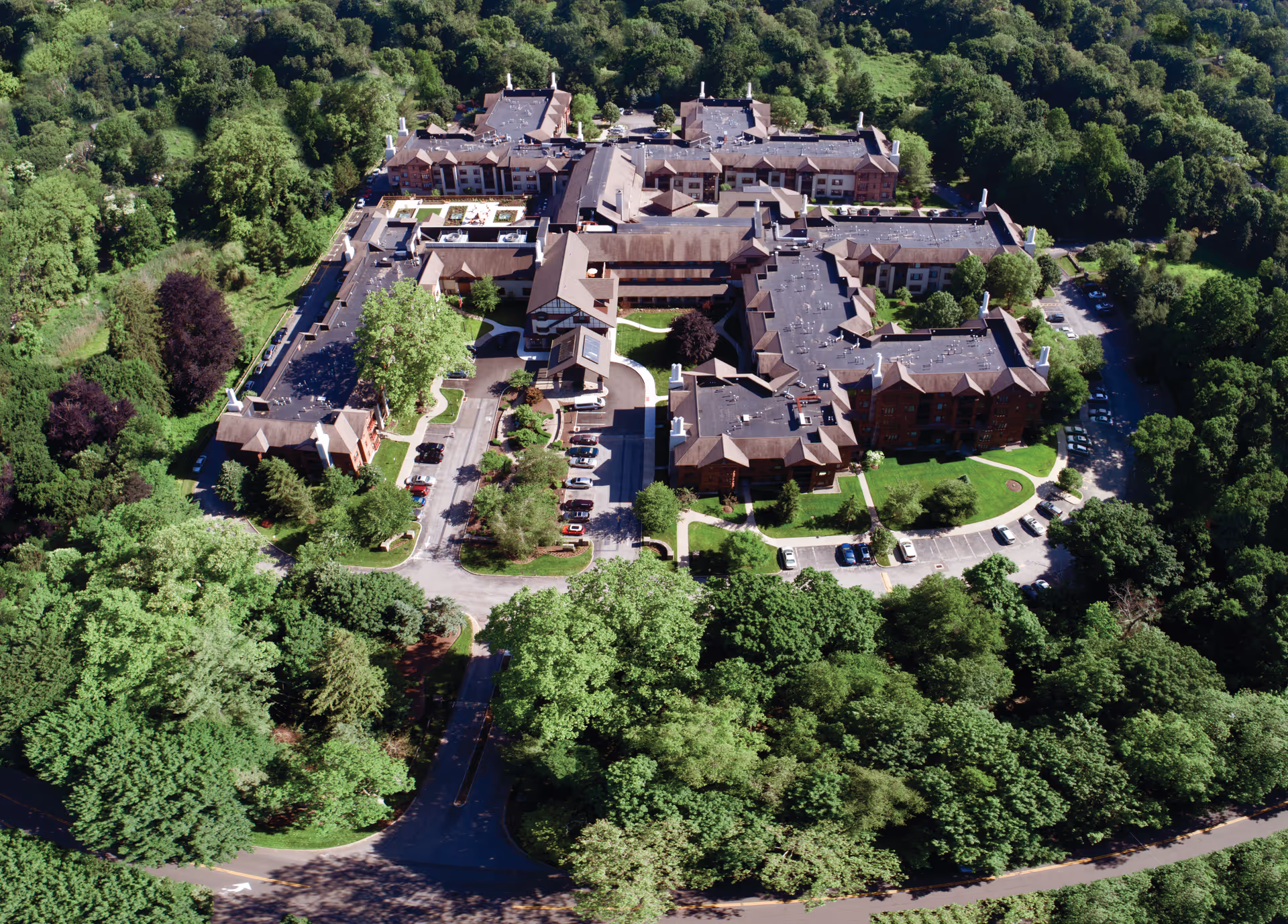 Aerial view of Edgehill Senior Living Community Stamford surrounded by dense green trees. The large multi-wing building has multiple parking areas and pathways around it, nestled in a lush, forested environment.