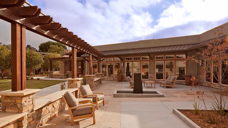 Outdoor courtyard area at Atria Valley View featuring cushioned wooden chairs, a central water feature, stone and wood pergolas, potted plants, and a building with large windows in the background under a partly cloudy sky.