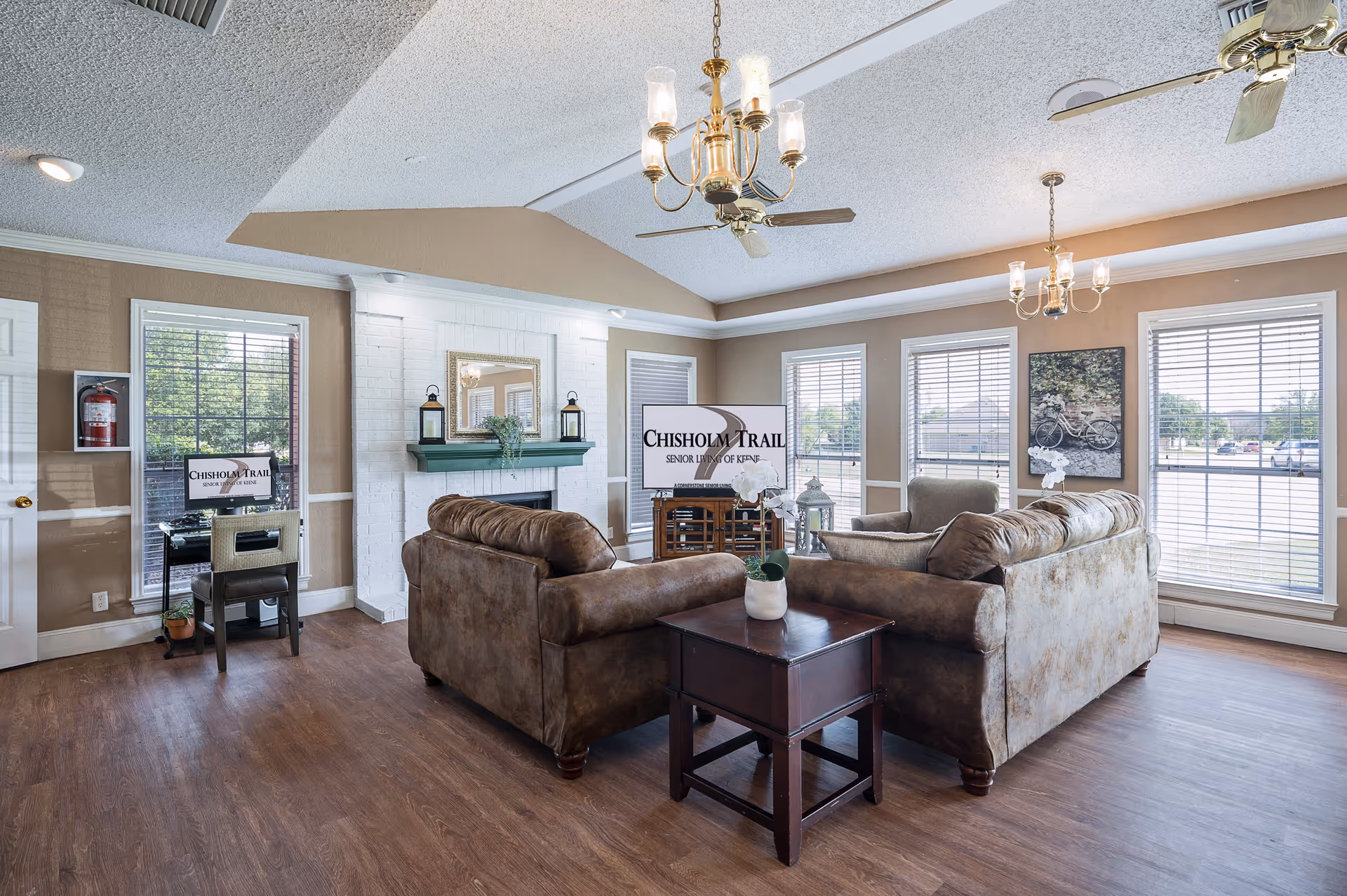 A bright and spacious living room in a senior living facility with two brown sofas arranged around a dark wooden coffee table. The room features large windows with white blinds, a white brick fireplace with a green mantel, and two chandeliers hanging from a vaulted ceiling with ceiling fans. There is a small desk with a chair and a computer screen displaying 'Chisholm Trail Senior Living of Kemp' near one window.
