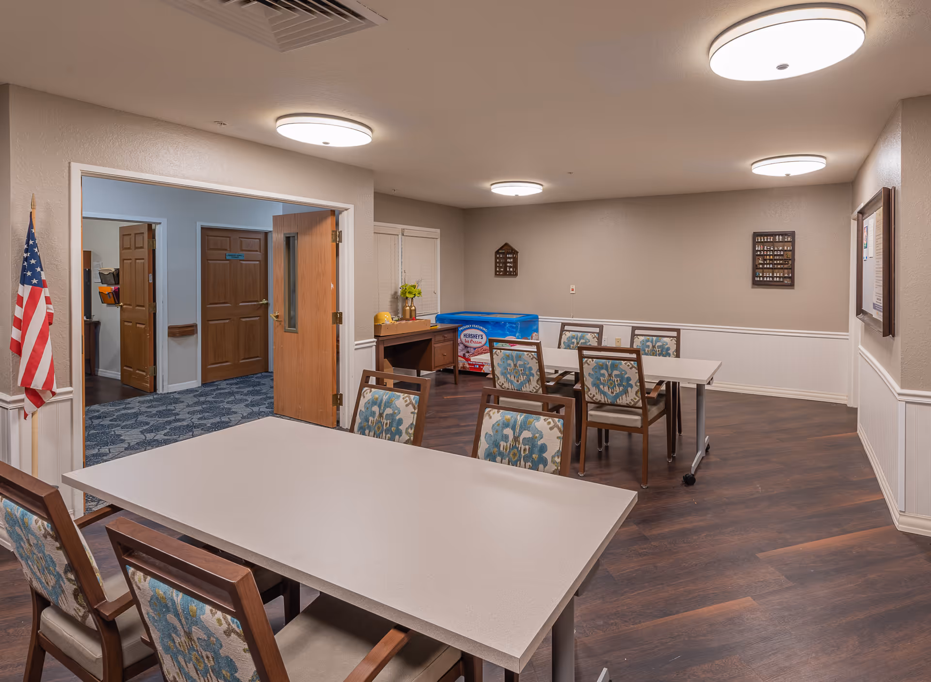 Interior room with several tables and chairs with floral upholstery, beige walls with white wainscoting, ceiling lights, an American flag near the entrance, and a blue Hershey's ice cream freezer against the far wall.