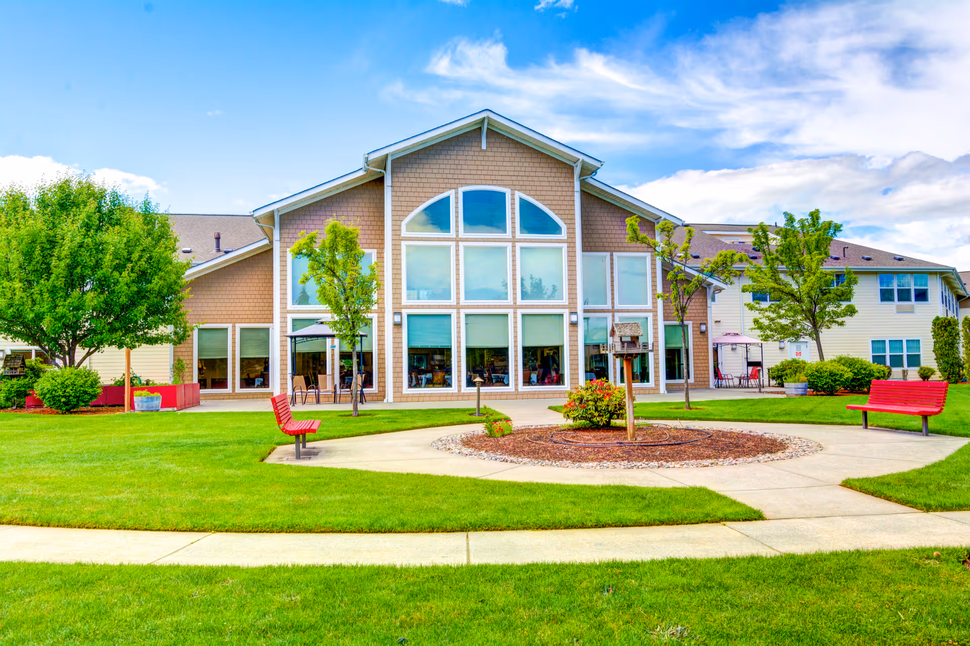 Exterior view of Hawks Ridge Assisted Living facility showing a large building with multiple windows, a well-maintained lawn, trees, red benches, and a circular paved area with landscaping under a partly cloudy blue sky.