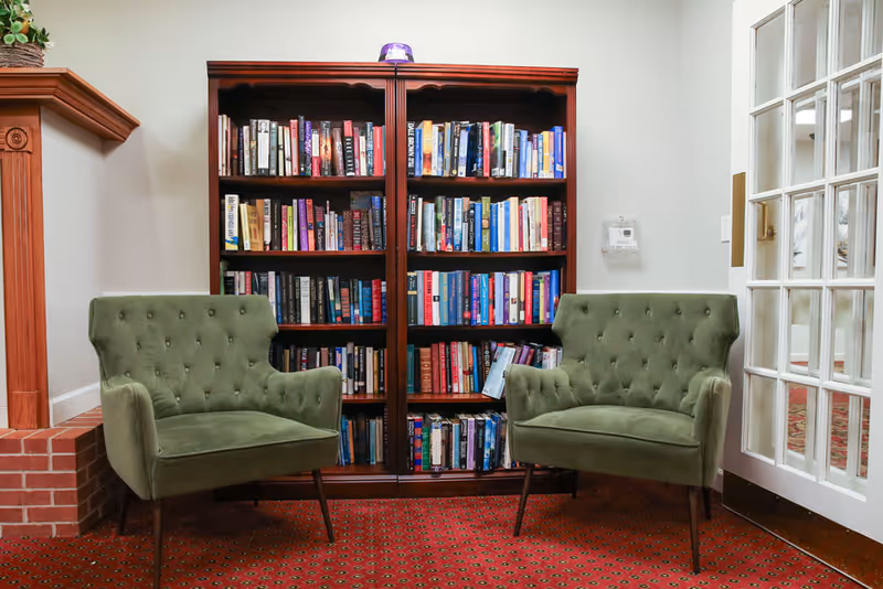 A cozy reading nook with two green upholstered armchairs facing each other, positioned on a red patterned carpet. Behind the chairs is a wooden bookshelf filled with various books. To the right is a glass-paneled door, and to the left is a brick fireplace mantel with a small plant on top.