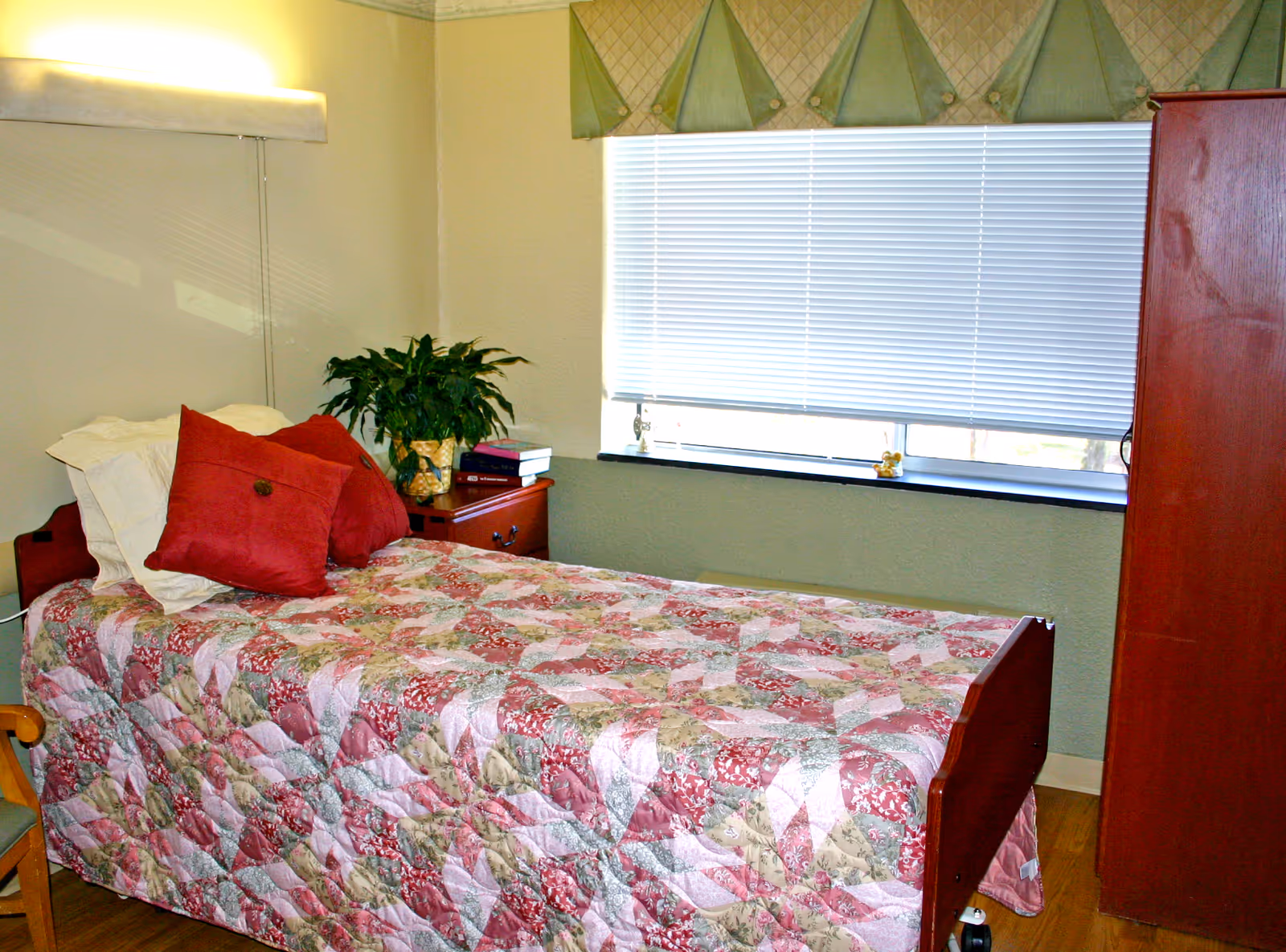 A single bed with a floral quilt and red pillows in a room with a wooden nightstand holding a plant and books, a window with closed blinds and a valance, and a wooden wardrobe.