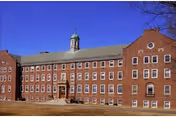 Exterior view of a large, multi-story brick building with many windows and a central entrance, topped with a small cupola under a clear blue sky.