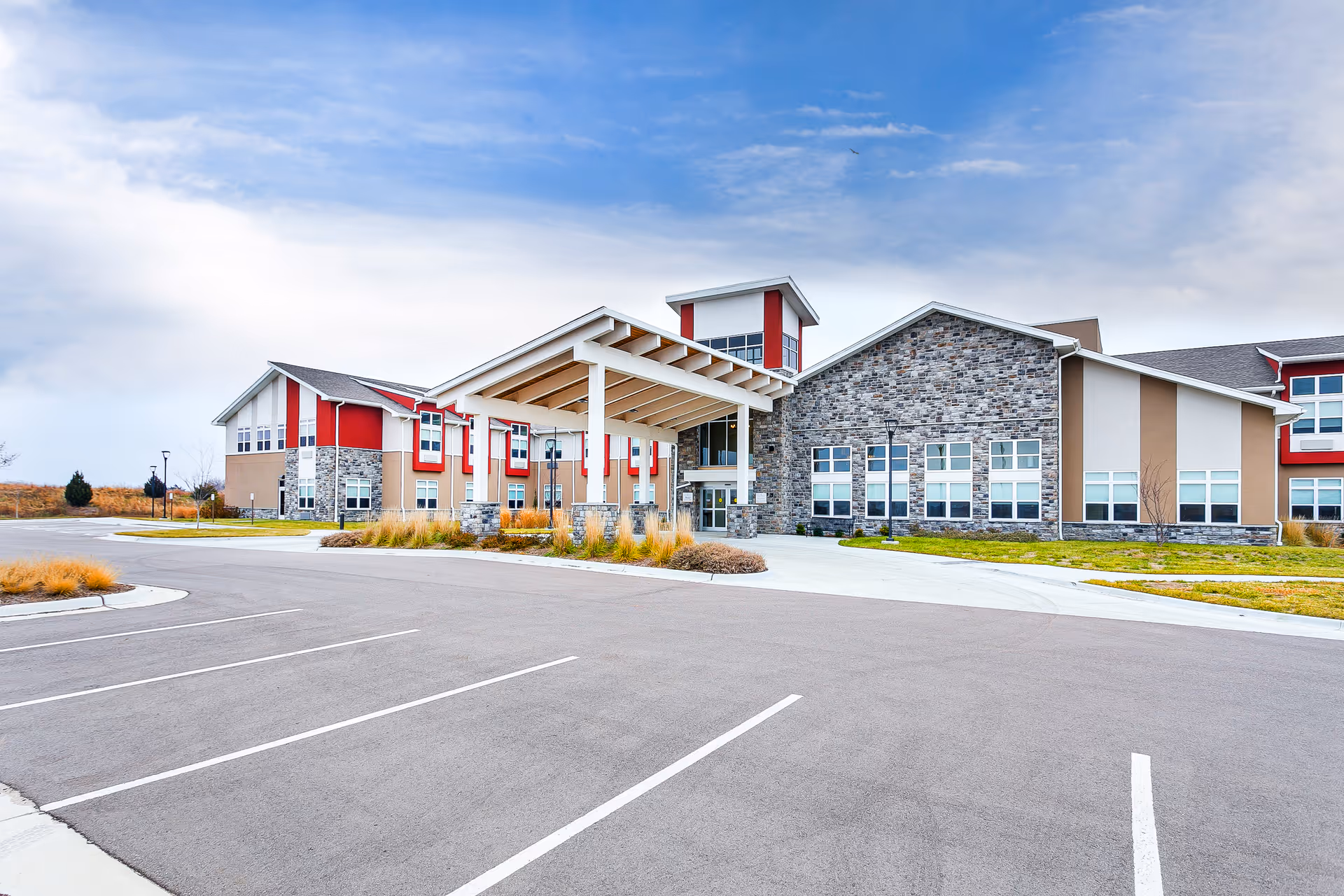 Exterior view of a modern senior living facility building with a covered entrance, stone and beige walls, red accents, and a large empty parking lot in front under a partly cloudy sky.