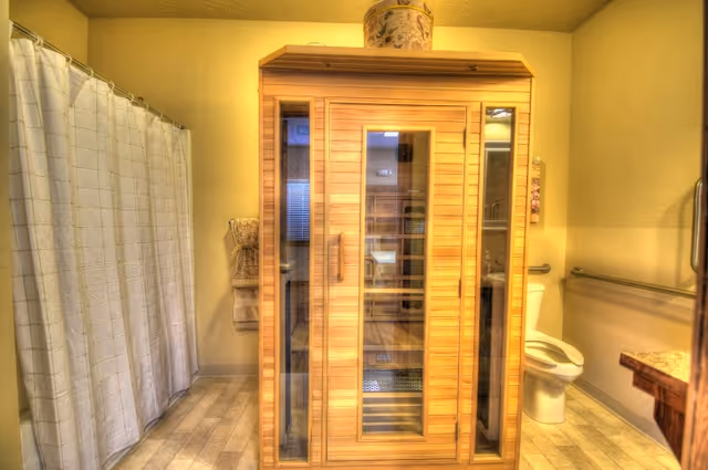 A bathroom featuring a wooden sauna with glass doors in the center. To the left, there is a shower area with a white curtain, and to the right, a toilet with a grab bar on the wall. The floor has light-colored tiles, and there are towels hanging on the wall next to the sauna.