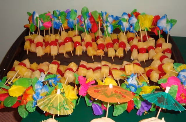 Tray of fruit skewers with pineapple chunks and maraschino cherries, decorated with colorful paper umbrellas and artificial tropical flowers.