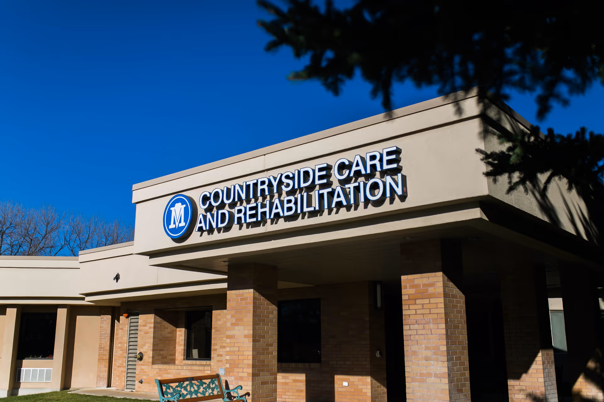 Exterior view of a building with a sign that reads 'Countryside Care and Rehabilitation' under a clear blue sky. The building has brick pillars and a beige facade with some trees partially visible in the foreground.