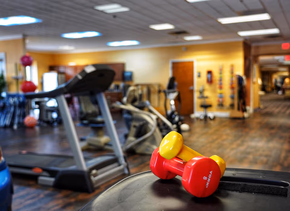 Interior view of a fitness room in a senior living facility with exercise equipment including treadmills, stationary bikes, and colorful dumbbells in the foreground. The room has wooden flooring and warm yellow walls with ceiling lights.